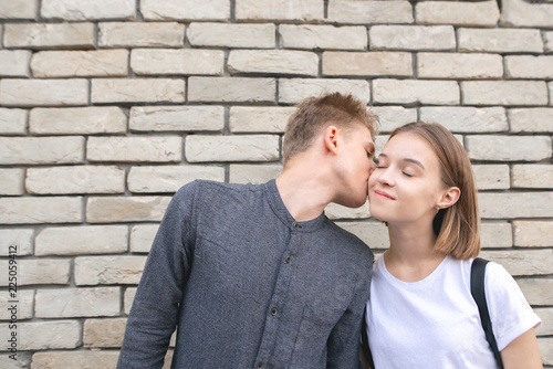 Happy smiling couple against the background of a brick wall, a young man kisses a girl on her cheek, she smiles. Love Story. Copyspace