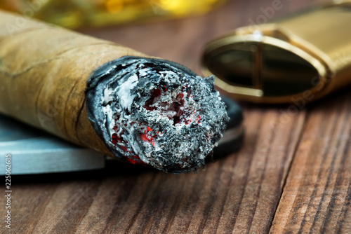 Smoldering cigar with ash on the end close-up next to a lighter and a piece of a glass of whiskey
