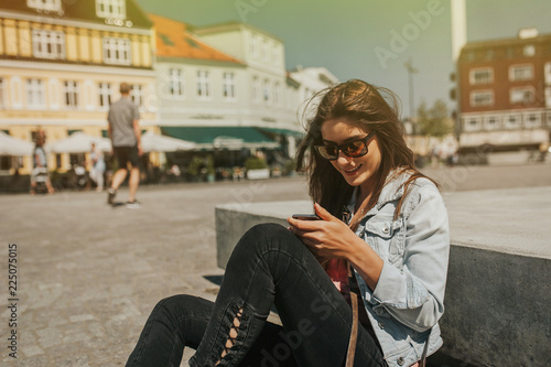 Young beautiful woman sitting in the city center holding a smartphone, smiling.