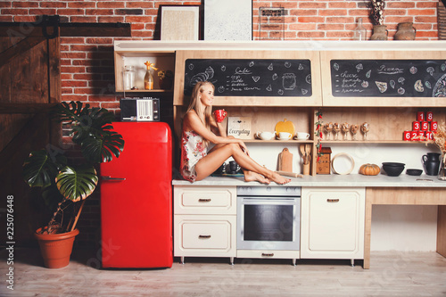 Pretty blonde long hair girl wearing pajama sitting on the comod, drinking her morning coffee and thinking about new day in the red loft comfortable sunny kitchen photo