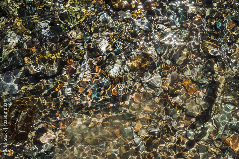 Varicolored detailed texture of stony bottom of transparent brook close up. Clean water in mountain creek. Colorful background of smooth stones in river stream with copy space.
