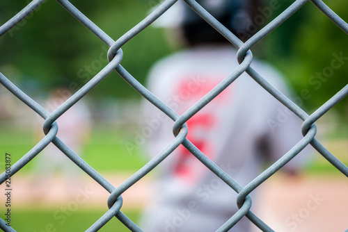 Unidentifiable youth junior league baseball player in team uniform, seen from behind through wire fence.
