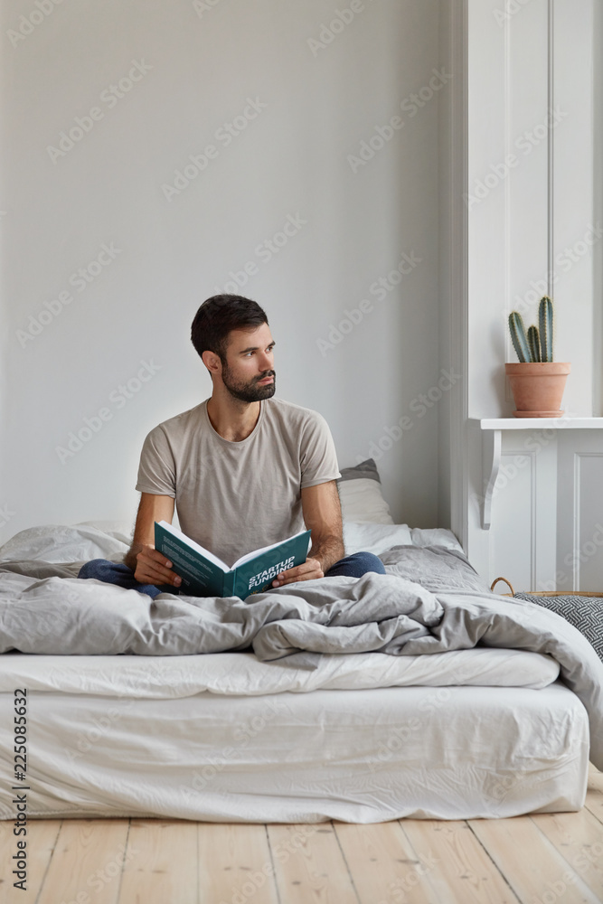 Attractive thoughtful bearded man with pensive expression, looks away, sits on bed, holds book, prepares for scientific research, reads necessary literature, busy with studying. Reading concept
