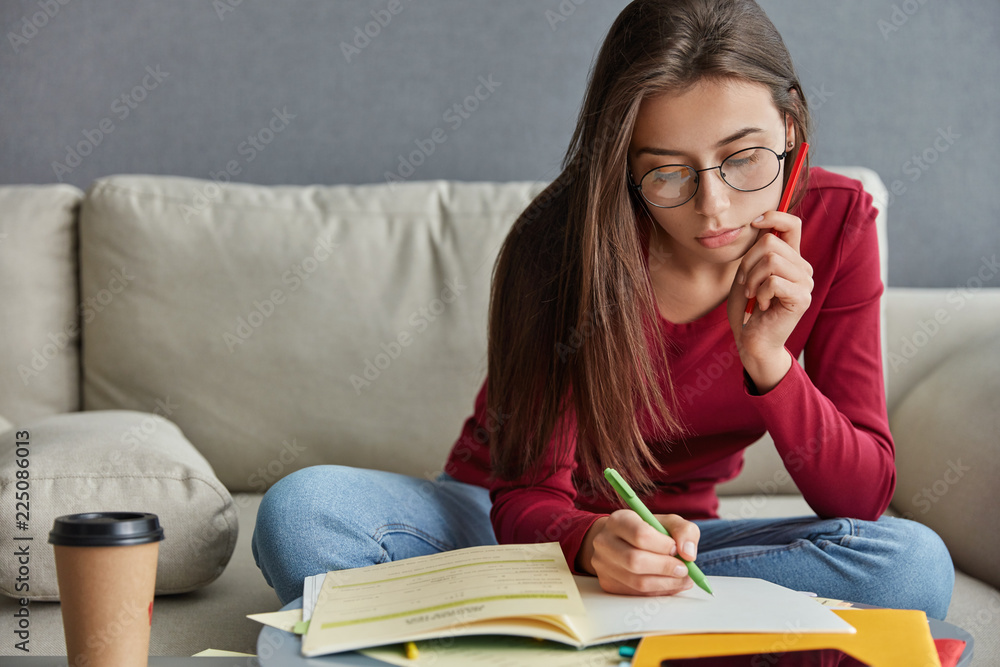 Shot of serious skilled student with pen in hand, writes curriculum vitae, wants to find extra job, busy with homework, dressed in casual clothes, drinks aromatic beverage, poses on couch in her room Shot of serious skilled student with pen in hand, writes curriculum vitae, wants to find extra job, busy with homework, dressed in casual clothes, drinks aromatic beverage, poses on couch in her room