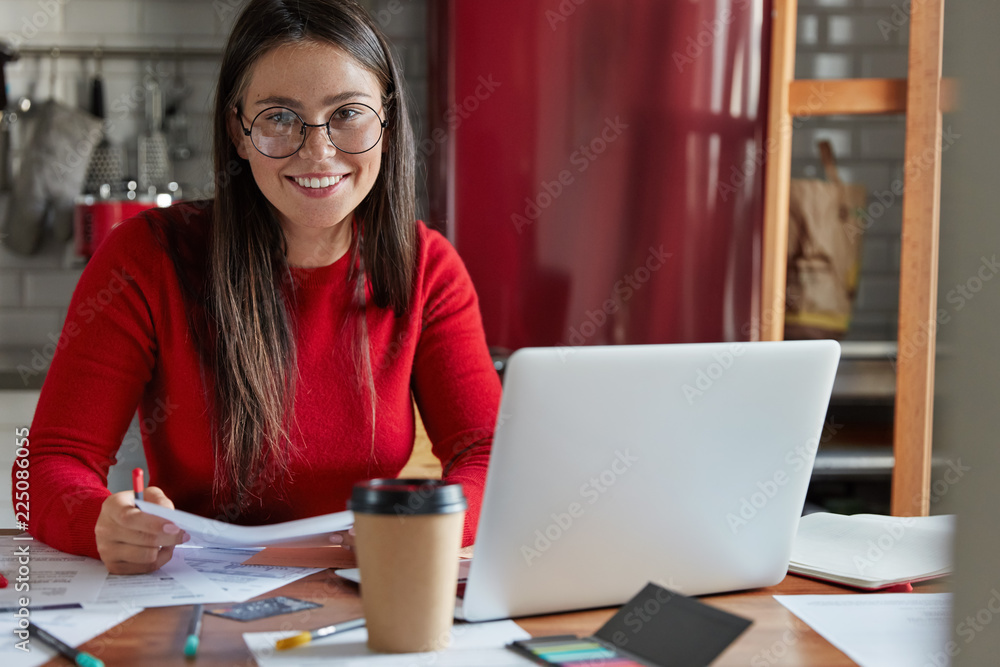 Glad student prepares for seminar, writes down homework checks text documentation, uses laptop computer, sits at kitchen table with cozy interior, enjoys takeaway coffee, checks delivered bills