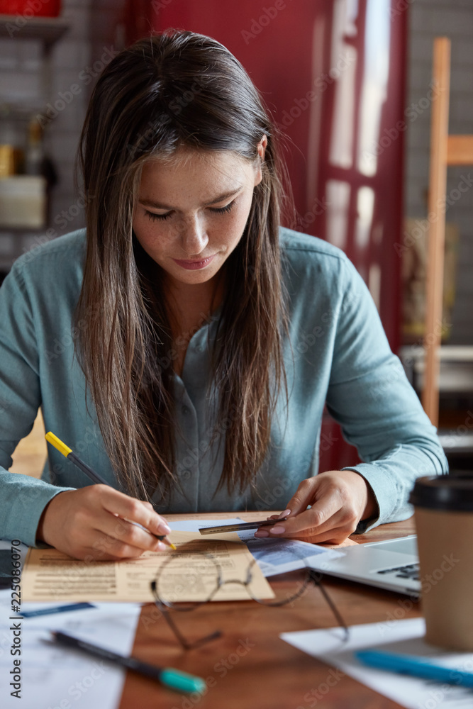Concetrated businesswoman sorts through papers, makes startup project, holds plastic cards, writes down number, dressed in shirt, poses over kitchen interior at home, has focused face expression Concetrated businesswoman sorts through papers, makes startup project, holds plastic cards, writes down number, dressed in shirt, poses over kitchen interior at home, has focused face expression