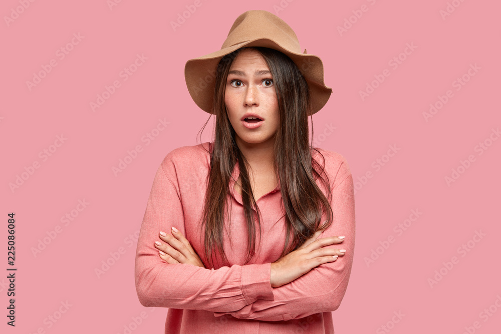 Waist up shot of indignant shocked woman keeps arms folded, looks with indignation, cant believe in shocking news, wears fashionable pink shirt and headgear, stands in studio alone. Emotions concept Waist up shot of indignant shocked woman keeps arms folded, looks with indignation, cant believe in shocking news, wears fashionable pink shirt and headgear, stands in studio alone. Emotions concept