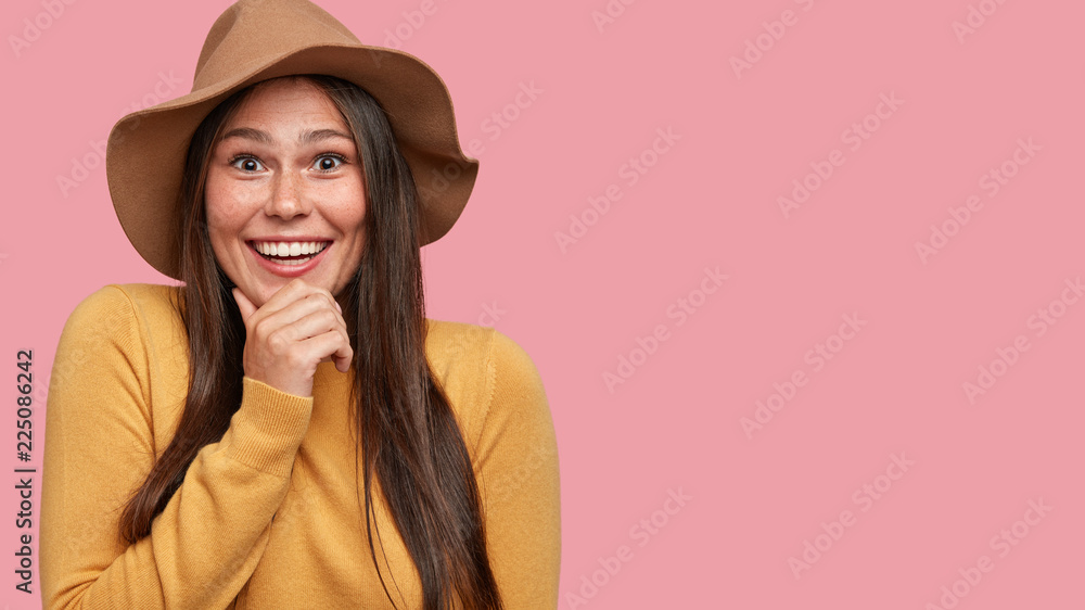 Photo of cheerful woman holds chin, has broad smile, being in high spirit as looks directly at camera, wears fashionable hat models against pink background with free space for your advertising content Photo of cheerful woman holds chin, has broad smile, being in high spirit as looks directly at camera, wears fashionable hat models against pink background with free space for your advertising content