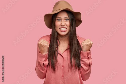 Photo of annoyed dissatisfied peevish girlfriend clenches teeth and fists, expresses anger as has quarrel with husband, cant control her emotions, dressed in brown hat, isolated on pink wall