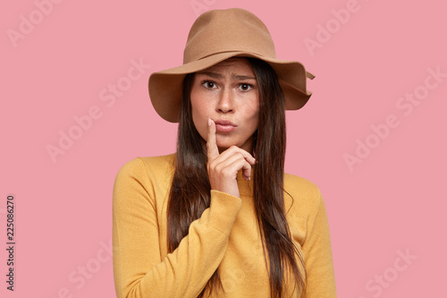 Displeased indignant teenager pouts lips, keeps finger near mouth, wears fashionable hat, casual sweater, look with puzzlement directly at camera, notices something unexpected, isolated on pink wall photo