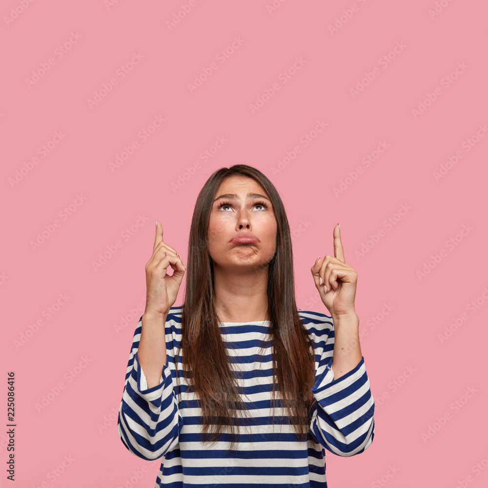 Photo of unhappy European girl being upset and dissatisfied with rainy weather, stands over pink background, dressed in oversized sweater, points with index fingers upwards. Awful advertisement Photo of unhappy European girl being upset and dissatisfied with rainy weather, stands over pink background, dressed in oversized sweater, points with index fingers upwards. Awful advertisement
