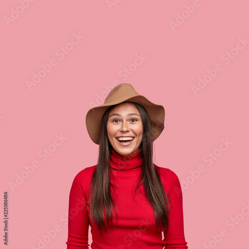 Vertical shot of good looking cheerful smiling girl has long straight hair, wears stylish hat and casual red turtleneck sweater, poses over pink background, free space for your advertising content.
