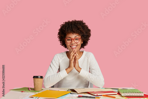 Positive young African American girl concentrated on course work, writes ideas, keeps hands together and smiles positively, happy to solve problems with project, poses against pink background photo