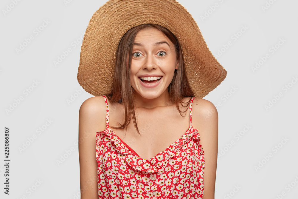 Emotional young lady has joyful expression, wears summer straw hat and fashionable dress, poses over white background, sees something very appealing. People, recreation and lifestyle concept Emotional young lady has joyful expression, wears summer straw hat and fashionable dress, poses over white background, sees something very appealing. People, recreation and lifestyle concept