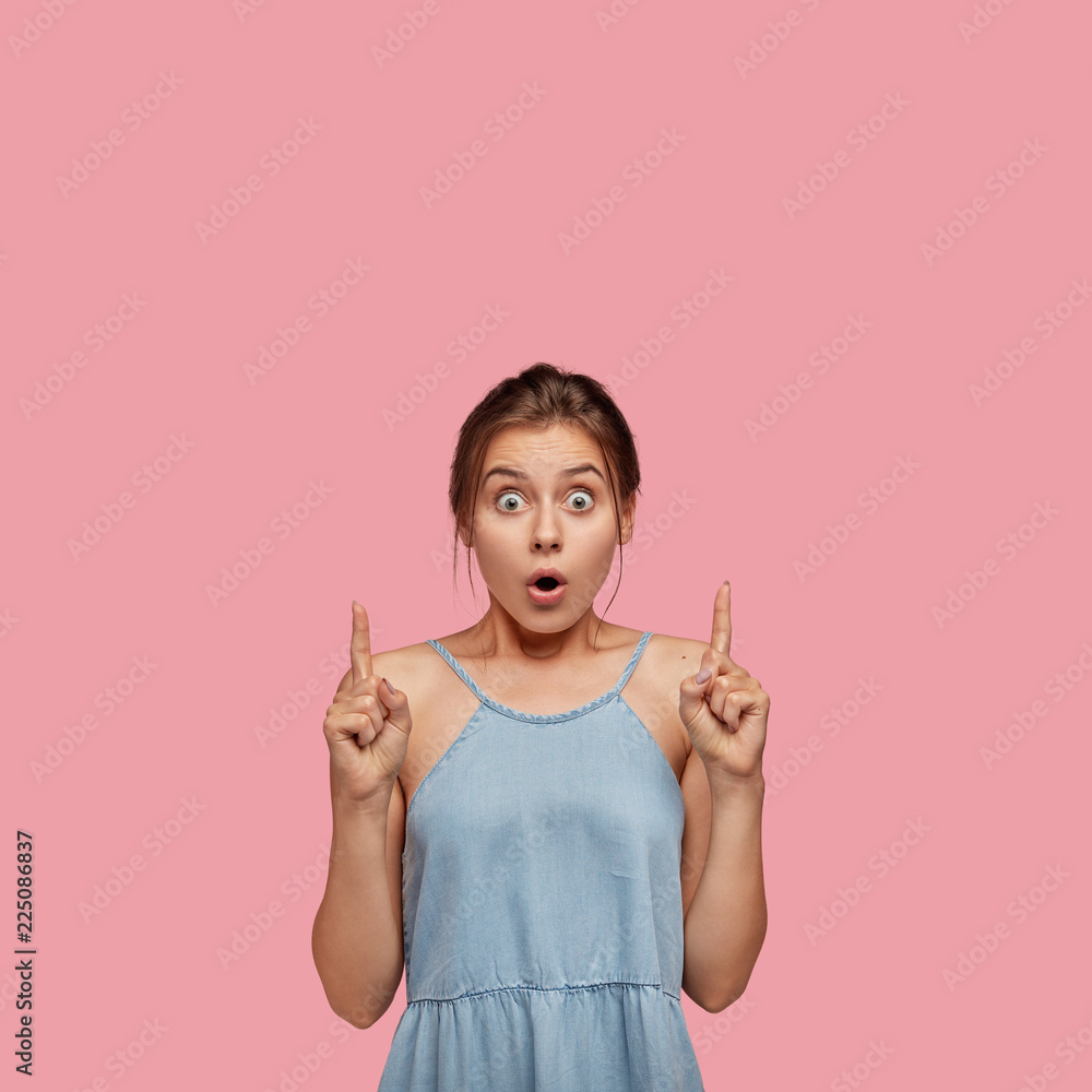 Indoor photo of stunned European girl with surprised expression, indicates with both index fingers upwards, shows something amazing, dressed in casual outfit, isolated on pink wall. Omg, look above Indoor photo of stunned European girl with surprised expression, indicates with both index fingers upwards, shows something amazing, dressed in casual outfit, isolated on pink wall. Omg, look above
