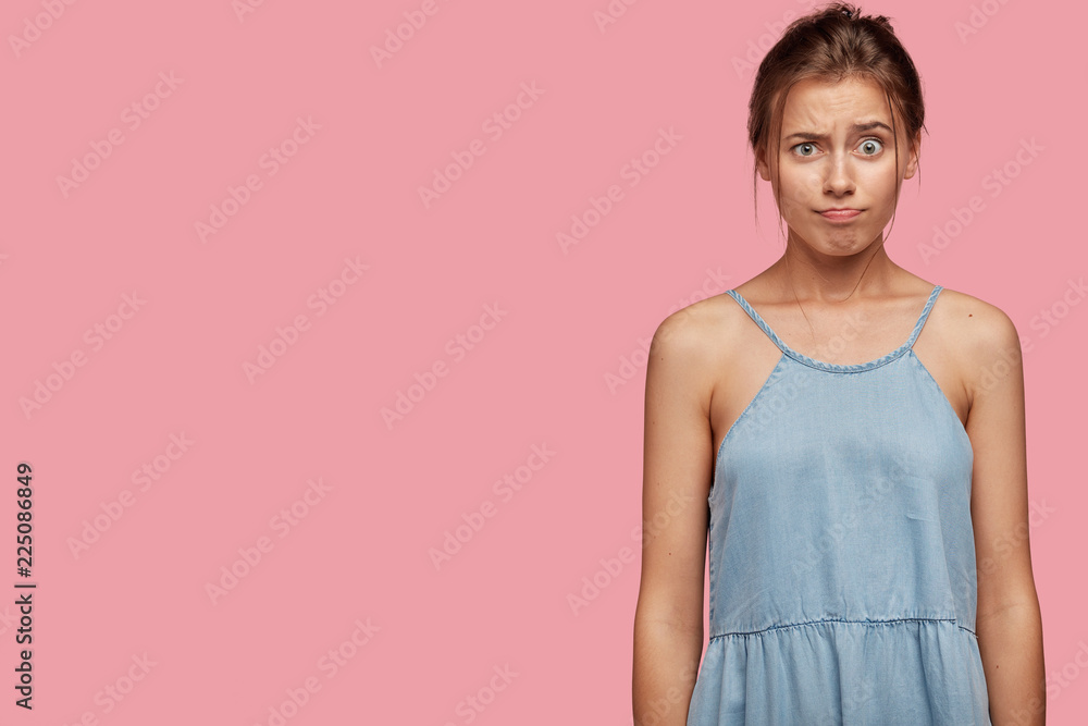 Dissatisfied cute teenager has annoyed facial expression, frowns face and purses lips, dressed in denim dress, models against pink background with free space for your promotional content or text Dissatisfied cute teenager has annoyed facial expression, frowns face and purses lips, dressed in denim dress, models against pink background with free space for your promotional content or text