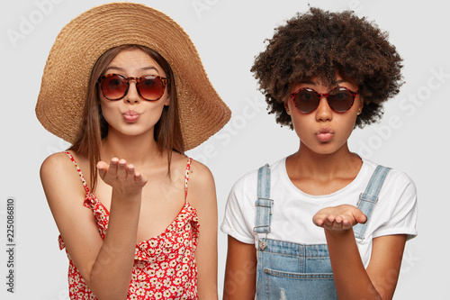 Two multiethnic sisters blow airkiss at camera, wears trendy shades, summer clothing, express love or say goodbye on distance, pose against white background. People, style and diversity concept photo
