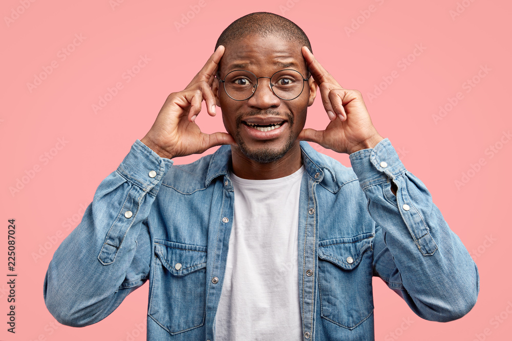 Studio shot of cheerful young black man keeps index fingers on temples, smiles positively, wears round spectacles, denim jacket, poses against pink background. Ethnicity and happiness concept Studio shot of cheerful young black man keeps index fingers on temples, smiles positively, wears round spectacles, denim jacket, poses against pink background. Ethnicity and happiness concept