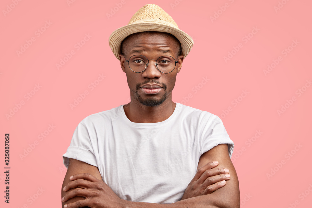 Indoor shot of sleepy black middle aged freelancer keeps hands crossed, feels fatigue, listens new information with bewilderment, wears round transparent glasses, isolated over pink background Indoor shot of sleepy black middle aged freelancer keeps hands crossed, feels fatigue, listens new information with bewilderment, wears round transparent glasses, isolated over pink background
