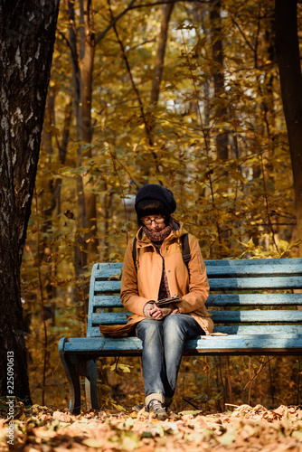 Young stylish hipster girl sitting on the bench in the autumn park and reading electronic book