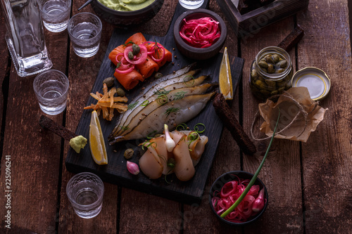 Assorted preserved fish on dark cutting board with vodka shots.