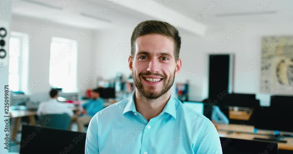 Portrait of young adult mid 30s handsome Caucasian male smiling and looking into camera, busy office in the background. 4K UHD 60 FPS SLOW MOTION Portrait of young adult mid 30s handsome Caucasian male smiling and looking into camera, busy office in the background. 4K UHD 60 FPS SLOW MOTION