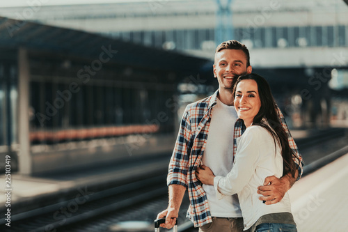 Portrait of beaming woman embracing positive male while standing on railway station during sunny day