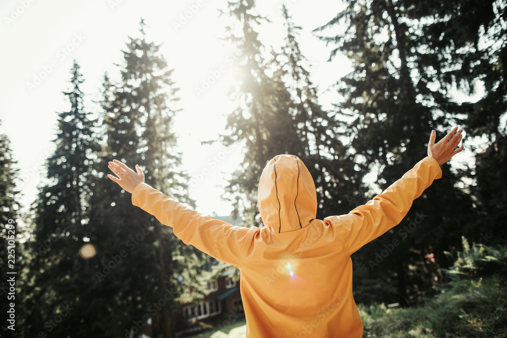 I am happy. Back view portrait of excited girl in yellow jacket with hood on her head raising hands in beautiful forest