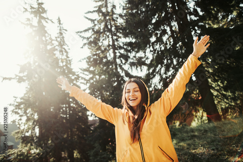 Wonderful day. Waist up portrait of happy girl in yellow jacket with hood on her head raising hands in coniferous wood. She is closing eyes in pleasure and smiling