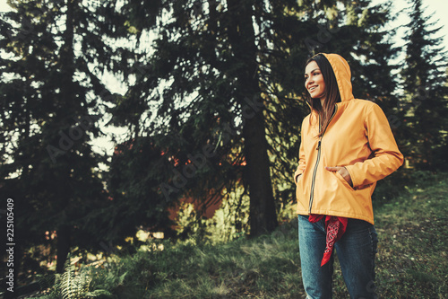 I am ready for adventures. Portrait of charming girl in yellow jacket enjoying sunny day in coniferous wood. She is looking away with smile and keeping hands in pockets