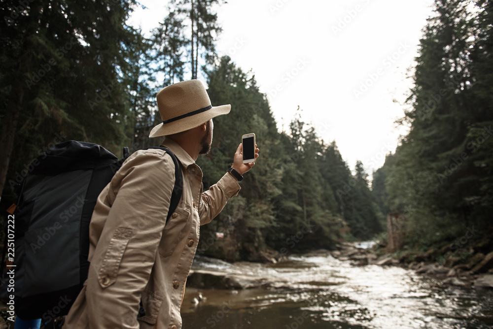 Taking photos. Active young traveler wearing a big hat and carrying heavy backpack while holding his modern smartphone and taking photos of the beautiful river Taking photos. Active young traveler wearing a big hat and carrying heavy backpack while holding his modern smartphone and taking photos of the beautiful river
