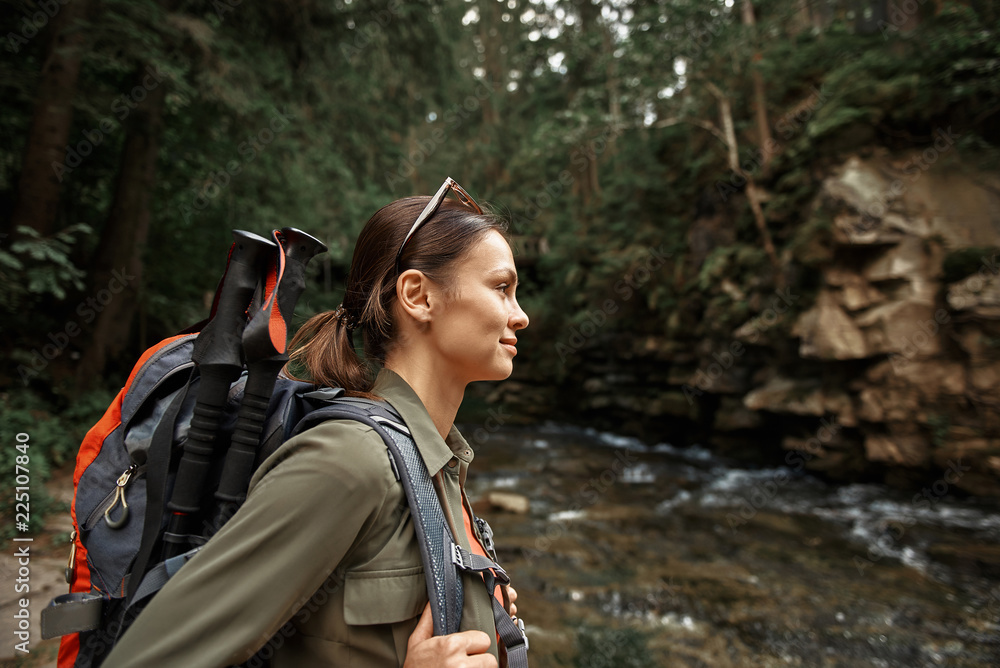 Active girl. Positive confident young woman smiling and enjoying her hiking trip while carrying heavy backpack with walking sticks