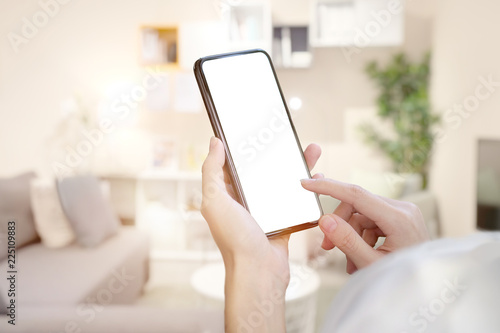 Female hand holding blank screen mobile smart phone over living room.