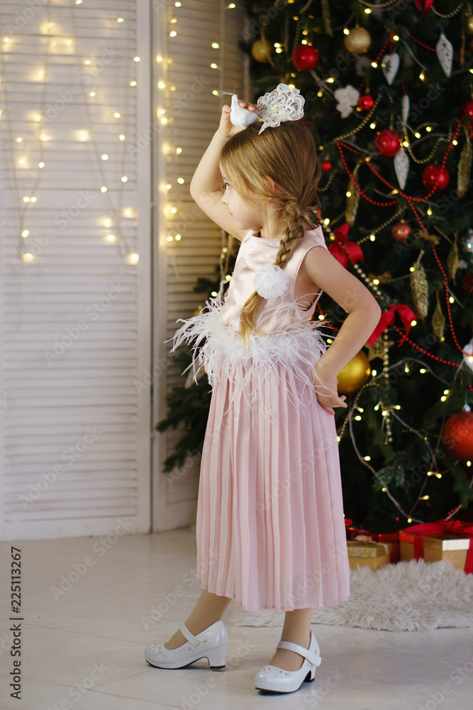 Little girl in a pink dress tries on decoration. Christmas tree with ornaments in the background. Happy childhood.