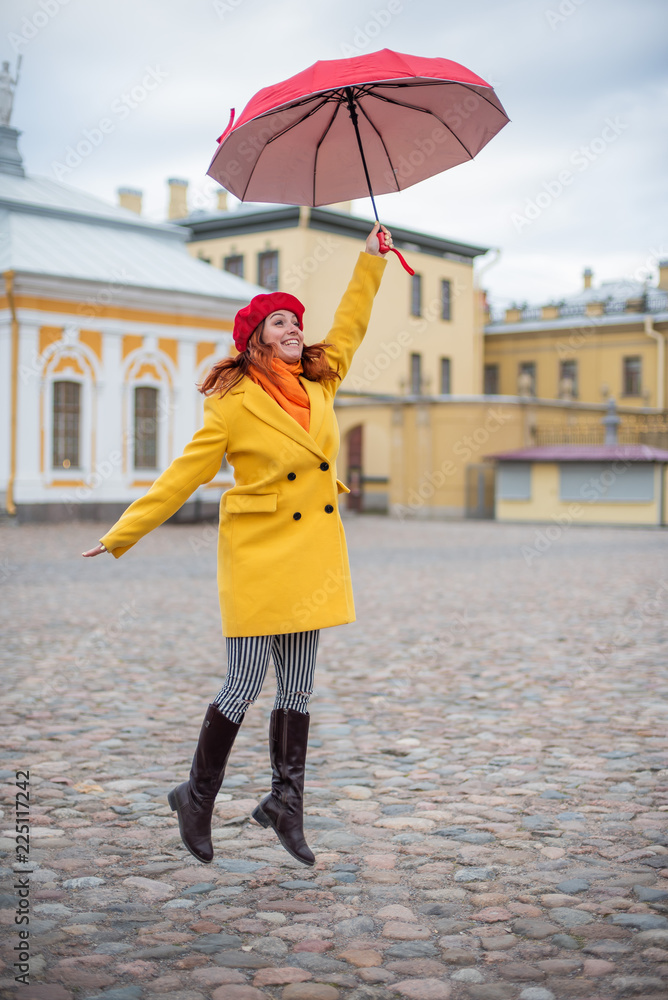 Fototapeta premium A red-haired woman in a yellow coat and a red beret is jumping with a red umbrella. A smiling beautiful woman in striped pants and a warm jacket flies with an umbrella, like Merry Poppins.