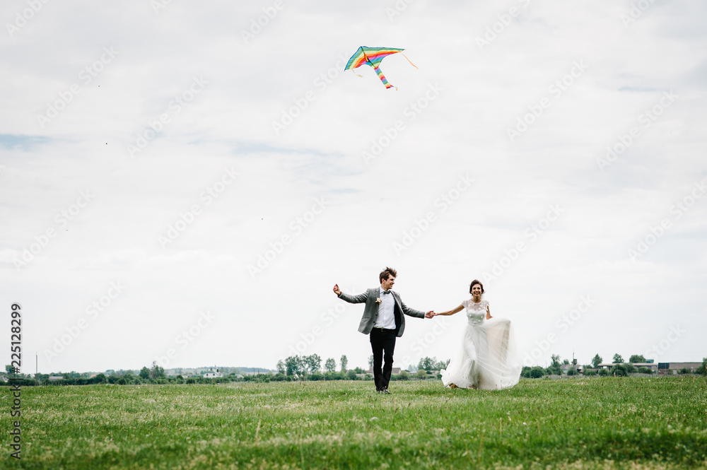 A wedding couple running around the field letting kite fly and play and hold the kite. Bride and groom walk near lake in the nature flying a kite together on a wedding day. A wedding couple running around the field letting kite fly and play and hold the kite. Bride and groom walk near lake in the nature flying a kite together on a wedding day.