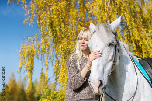 Pretty young blonde woman with a white horse in autumn day