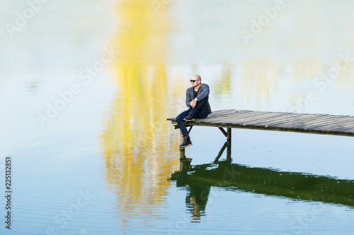 The bald young man in black sunglasses sit on the wooden pier