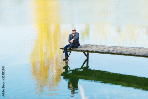 The bald young man in black sunglasses sit on the pier near the lake