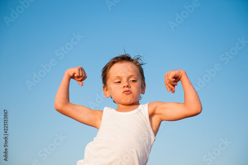 Canvas Print strong healthy little boy in shirt showing his muscles on blue sky background