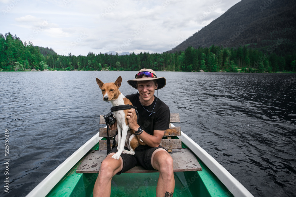 Happy smiling young man, sits in boat with basenji puppy or dog on lap in middle of alpine beautiful lake. Concept wanderlust and travel destination goals, active healthy lifestyle with pets