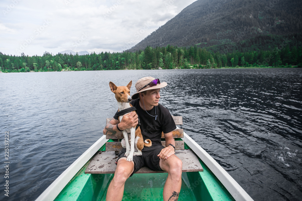 Urban traveller and adventurer in panama hat with authentic tattoos sits in rowing boat with best buddy friend, basenji dog, camping and hiking exploring world and nature active lifestyle together