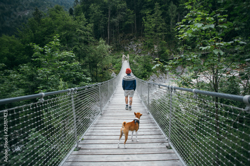 Hiker, traveller or tourist on adventure hike or exploring trip walks in middle of hanging wooden bridge in mountain forest or river, wears blue wool sweater, boots and beanie, with brown basenji dog