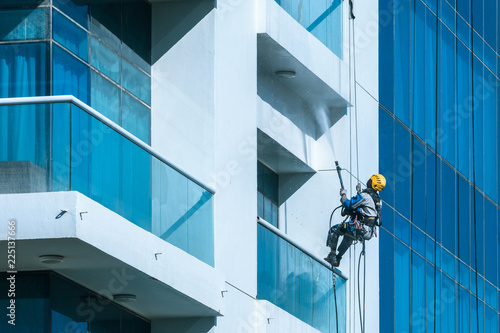 Worker wearing safety harness wash glass facade at height on modern high rise building. Professional rope access