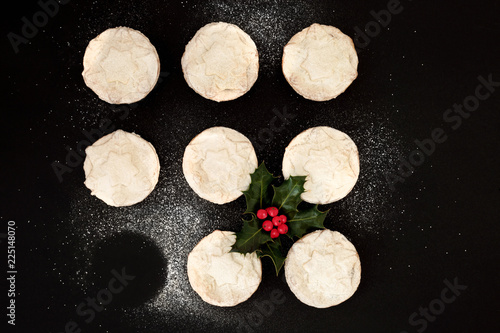 Delicious freshly baked Christmas mince pies with one missing, with holly berry leaf sprig and icing sugar dusting on black background. Top view.