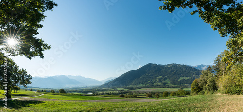 panorama countryside view with a road leading through fields and forest with the sun shining and mountains behind