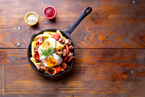 CHILEAN FOOD. CHORRILLANA - french fries topped with beef sliced, tipical sausages chorrisos, fried onion and eggs served in cast iron pan with sauces. Wooden background. top view photo
