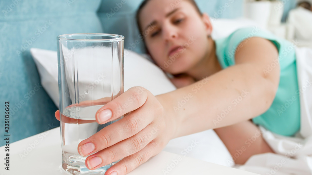 Closeup image of young woman feeling unwell taking glass of water from bedside table Closeup image of young woman feeling unwell taking glass of water from bedside table