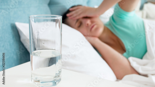 Closeup image of glass of water against woman with headache lying in bed