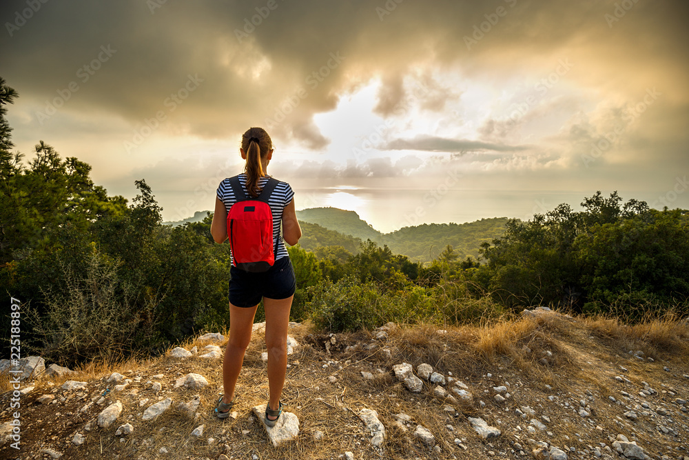 A young girl tourist with a backpack stands on the edge of a mountain and looking into the distance at dawn or sunset